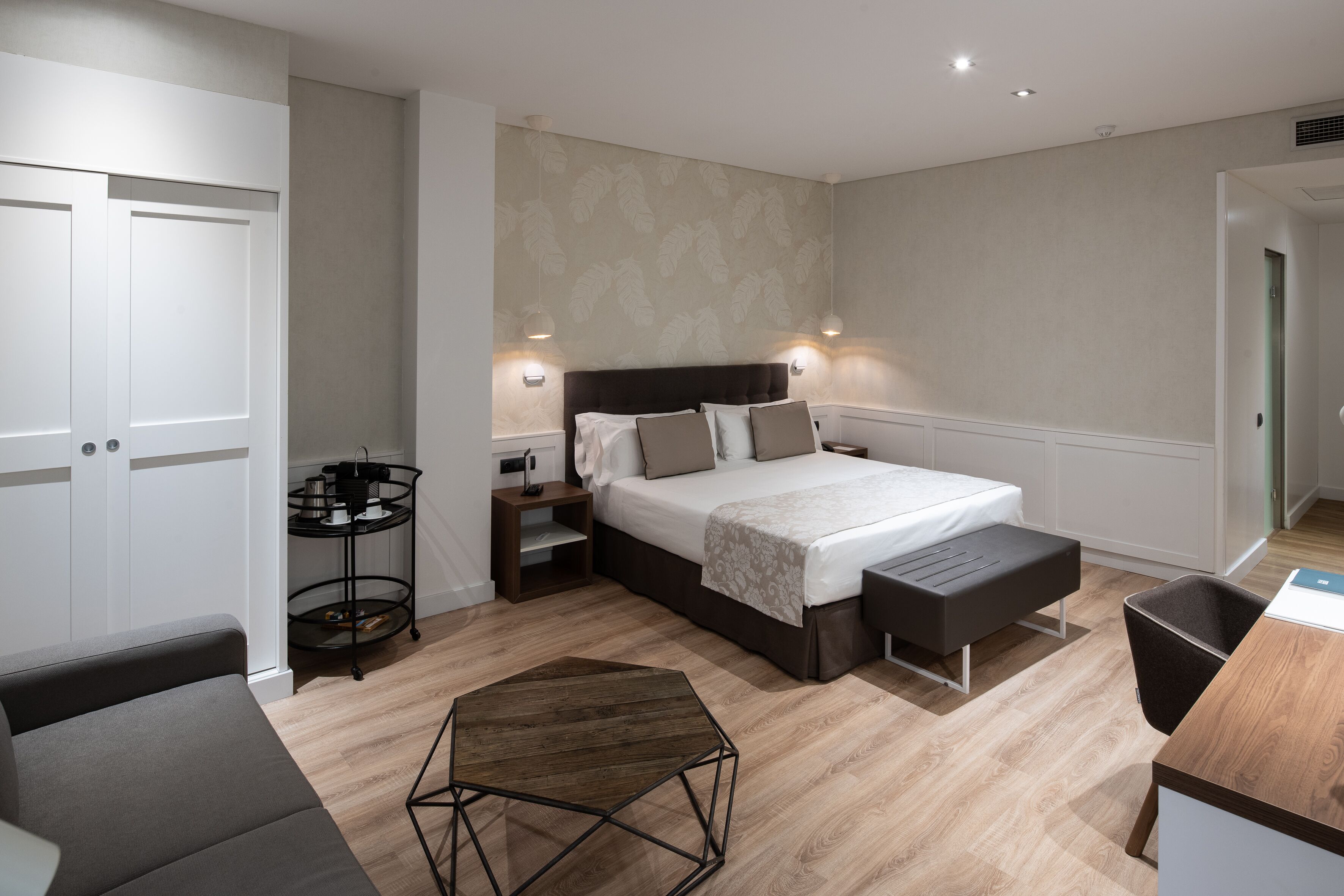 Une chambre à coucher moderne avec un lit, des tables de chevet, une petite table ronde et un banc.