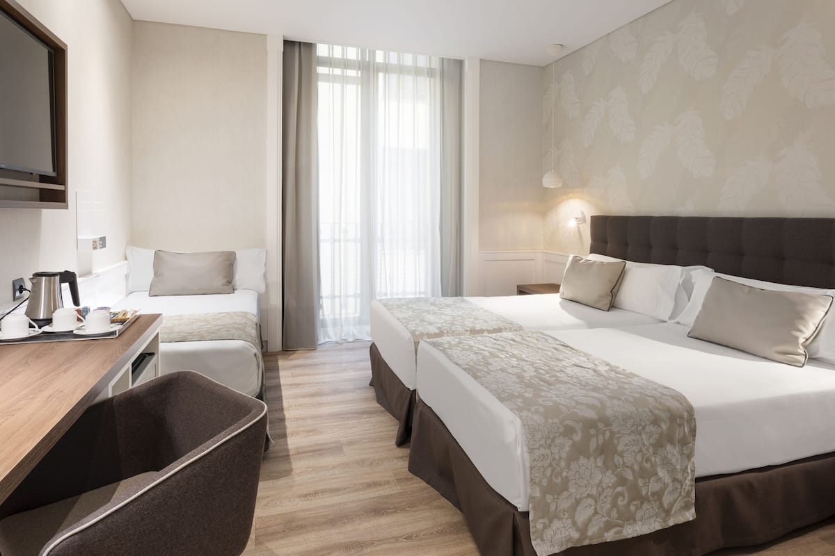 A hotel room with two beds, a desk with a chair, a television, and a large window with curtains.