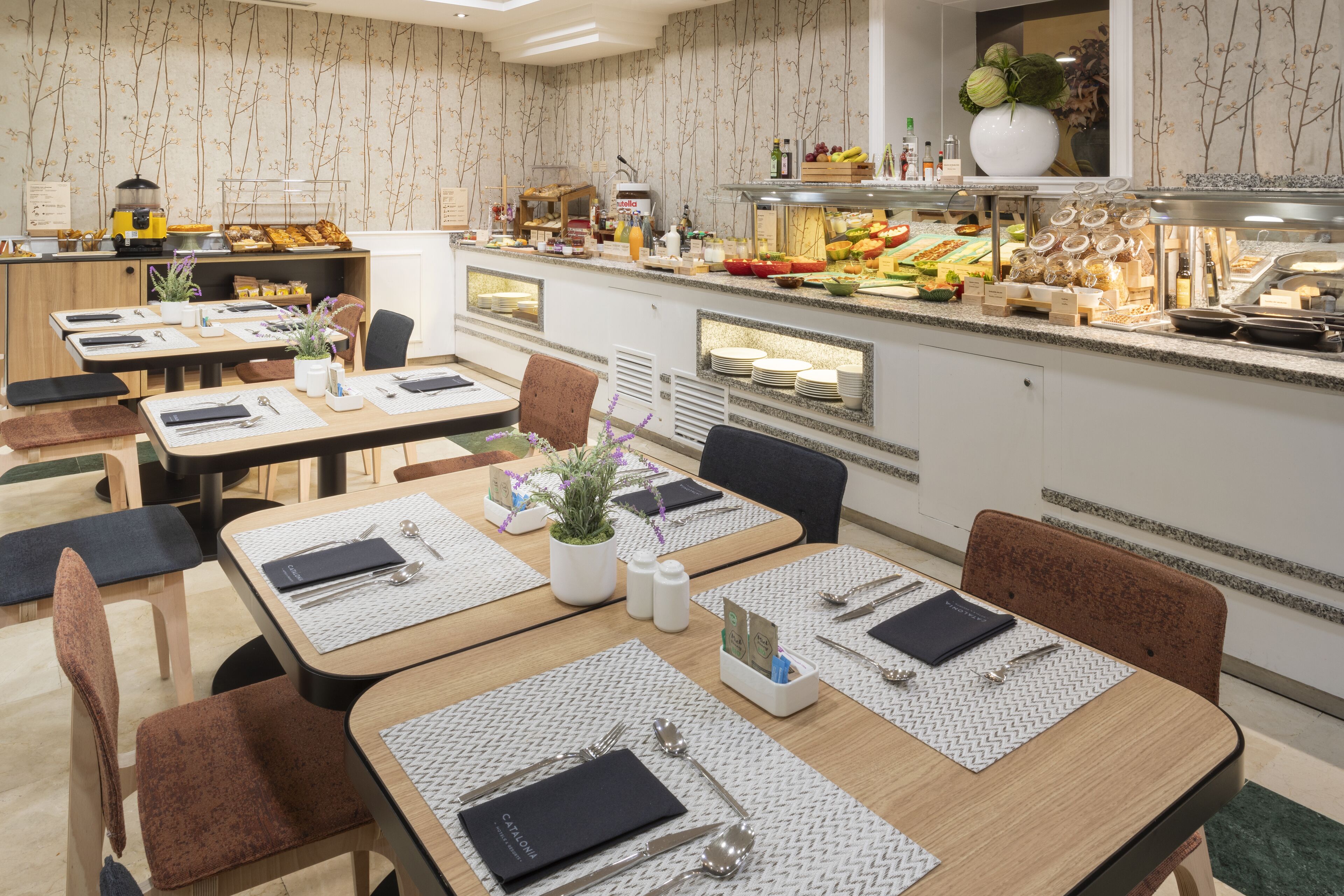 A restaurant with multiple dining tables set with placemats, cutlery, and menus. There is a buffet area with various food items and a display of condiments.