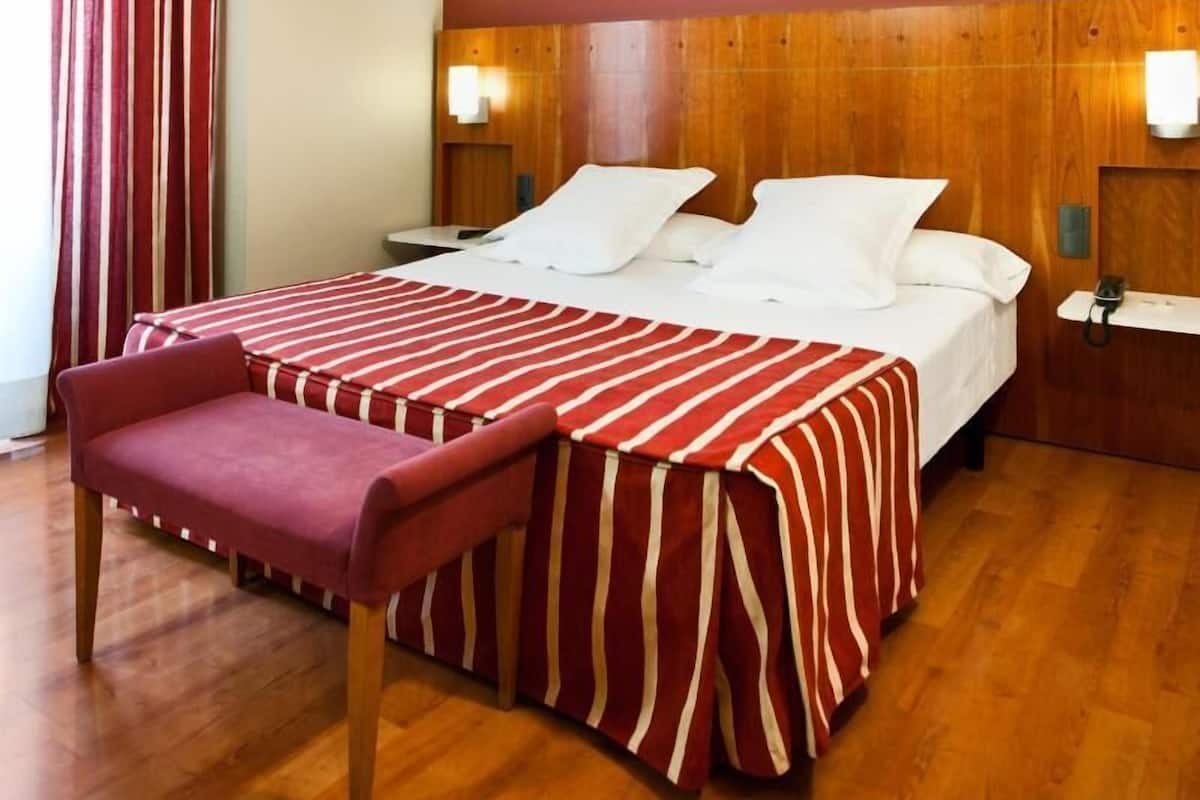 A hotel room with a bed, a wooden headboard, a bedside table, a chair, and a window with curtains.