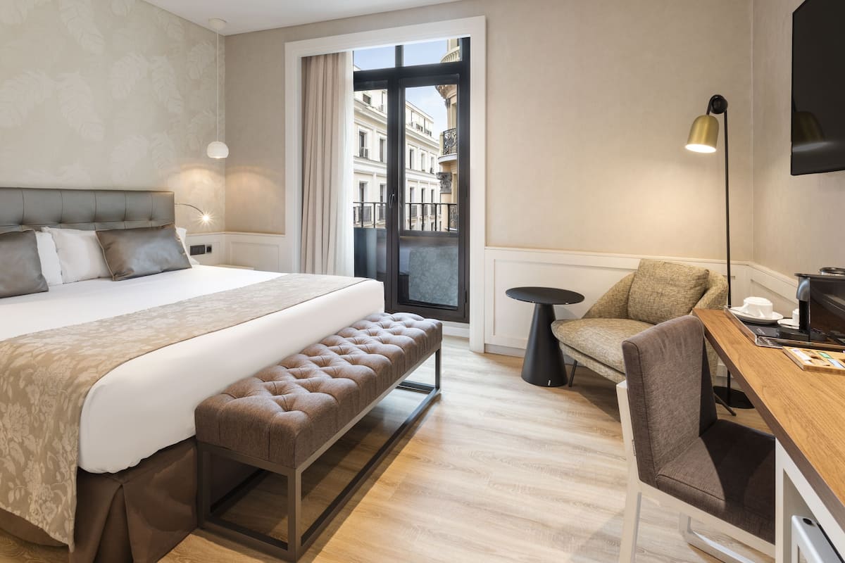 A hotel room with a large bed, a bench, a desk, and a view of the city through a balcony.