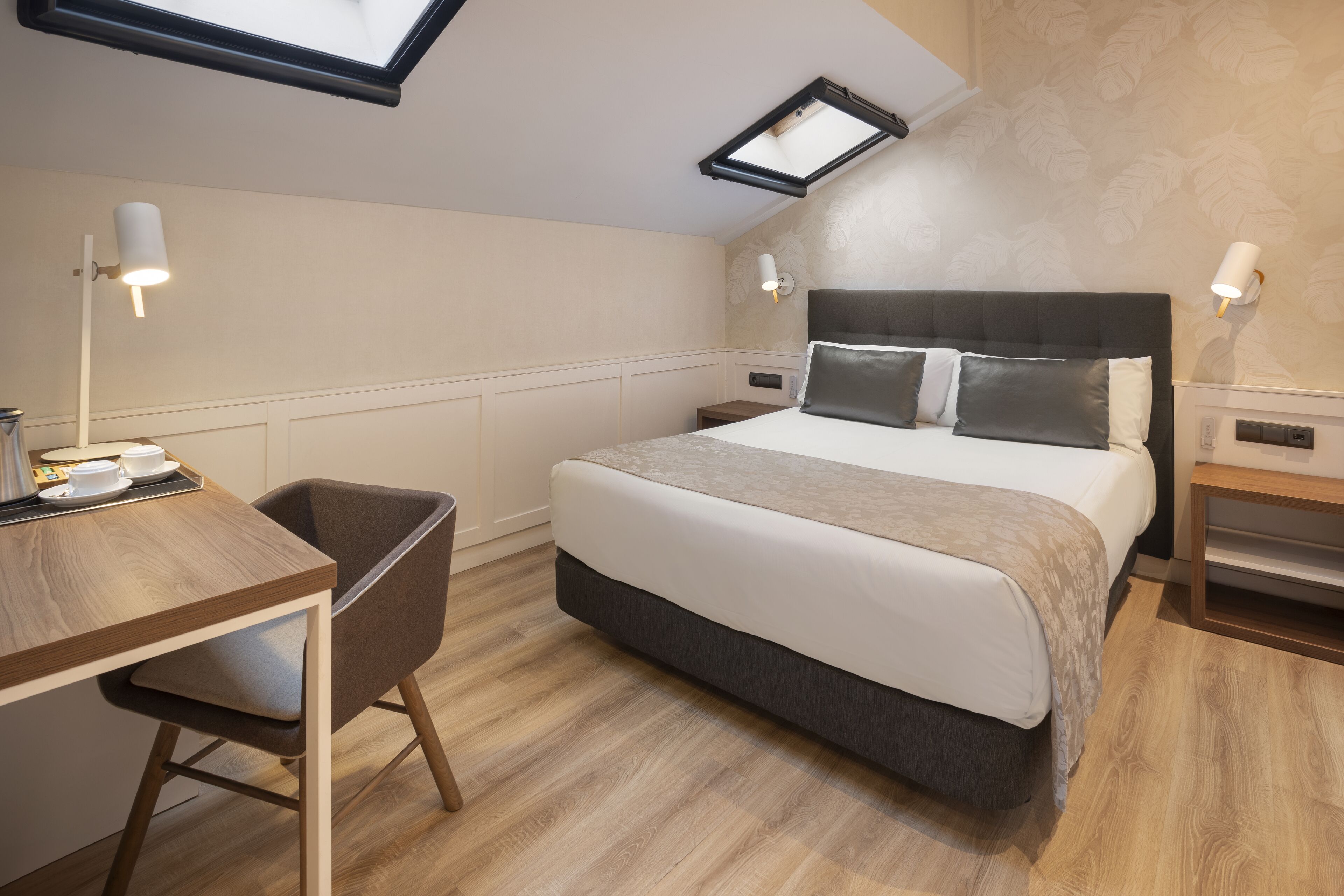 A modern hotel room with a large bed, a desk, and a chair. There is a skylight above the bed and wall-mounted lamps.
