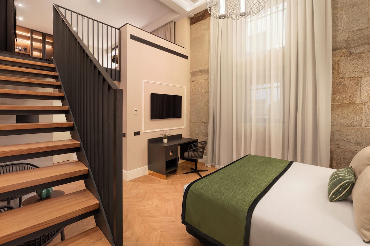 A modern bedroom with a wooden staircase, a desk with a chair, a bed with a green blanket, and a TV mounted on the wall.