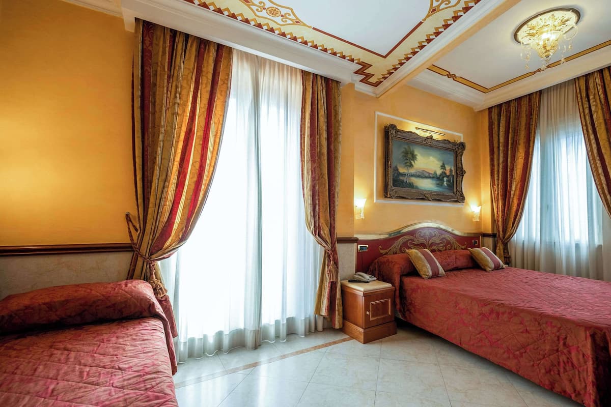 A hotel room with two beds, a nightstand, a painting on the wall, and a chandelier.
