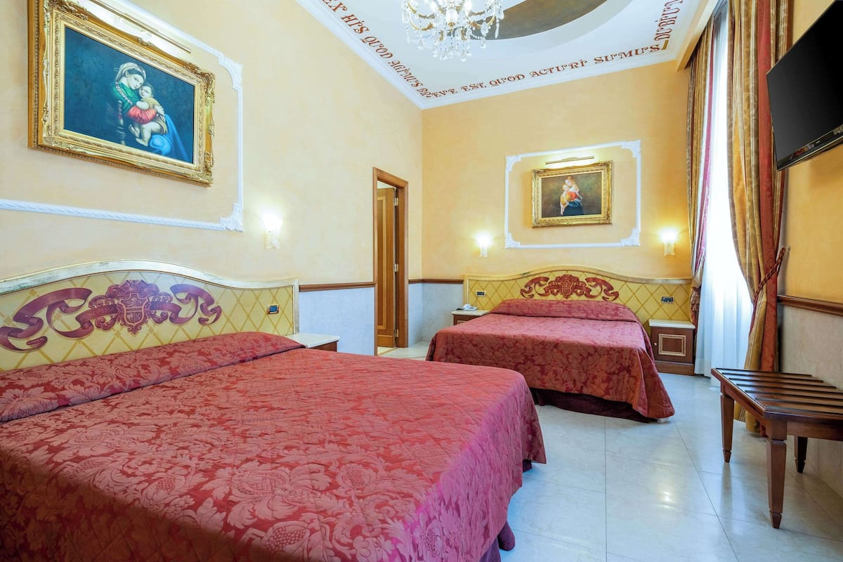A hotel room with two beds, a chandelier, and two framed paintings on the walls.