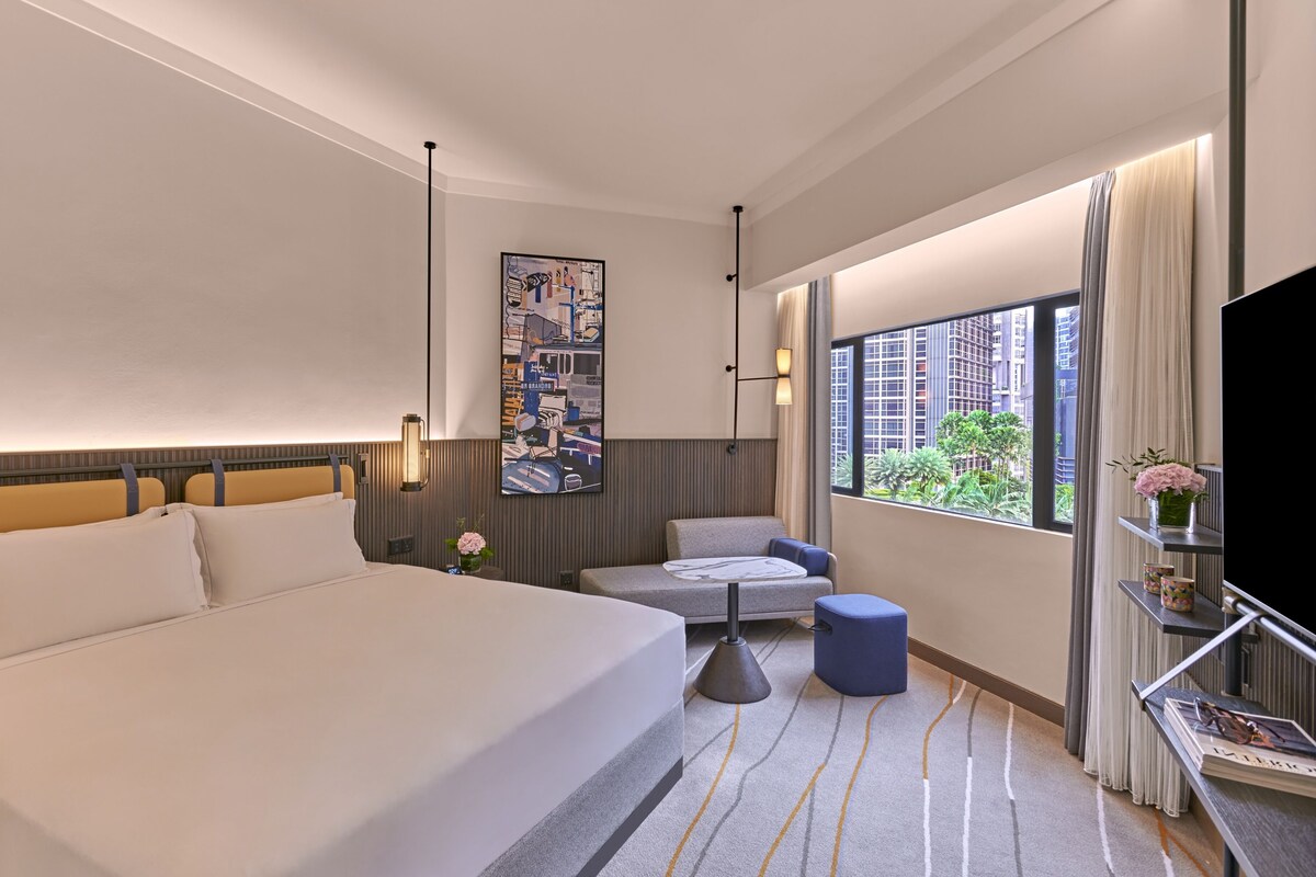 Habitación de hotel con una cama grande, un sofá, una mesita y un televisor. Se ve un ventanal con vistas a edificios y vegetación.
