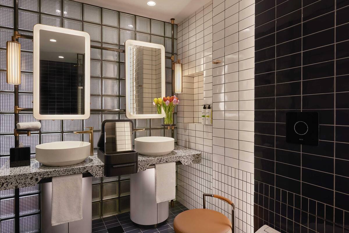 Een moderne badkamer met twee wastafels, een spiegel, een handdoekrek, een stoel en een bedieningspaneel aan de muur.