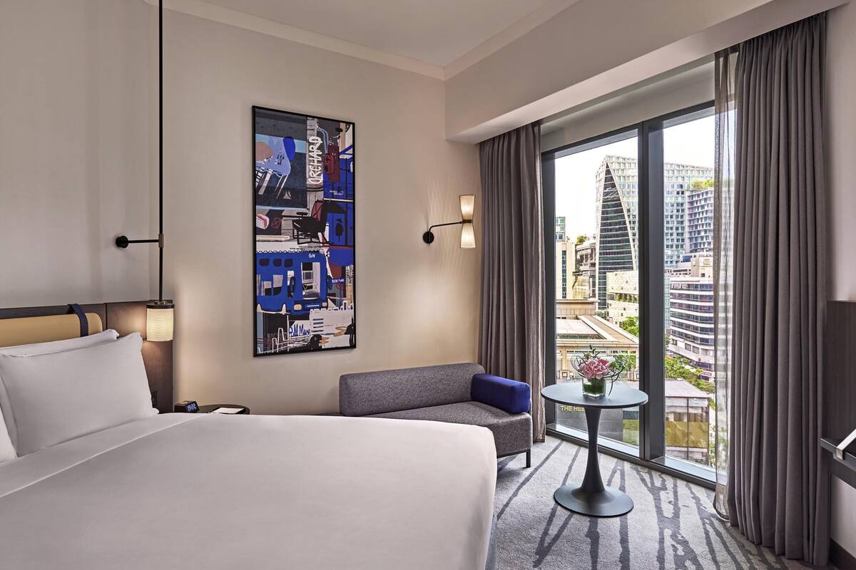 Una habitación de hotel con una cama grande, un sofá, una mesita y una obra de arte con un motivo urbano en la pared, además de vistas a edificios altos por la ventana.