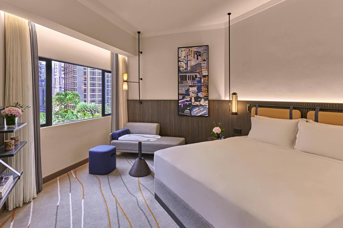Un cuarto de hotel con cama, una silla, una mesita, una estantería, un cuadro y una ventana que ofrece vistas a edificios y zonas verdes.