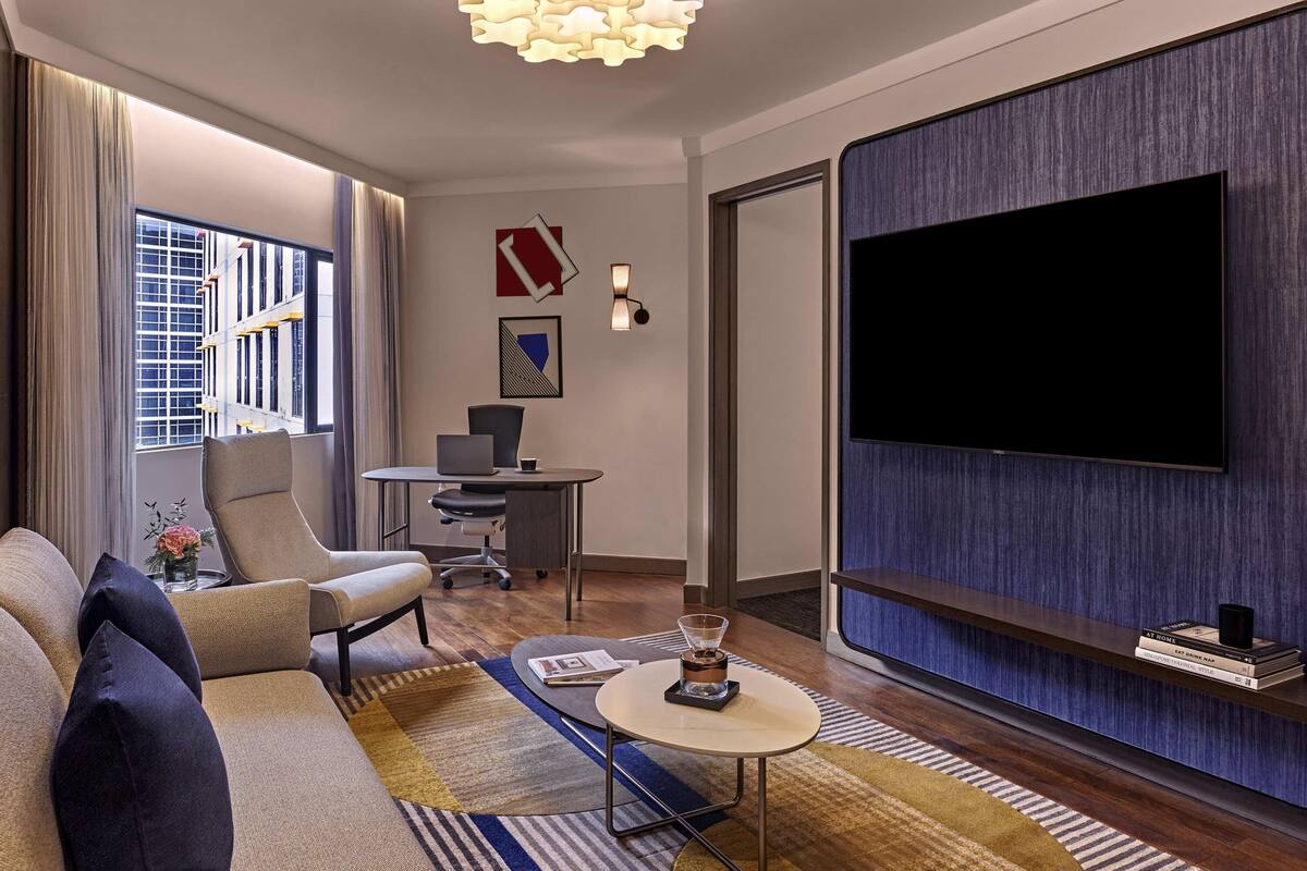 Una habitación de hotel moderna con sofá, escritorio con laptop, televisión y un ventanal grande.