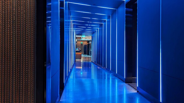 Un couloir moderne, éclairé de lumière bleue, avec un sol réfléchissant, qui mène à un espace d’accueil.