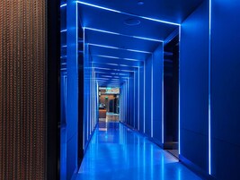 Un pasillo moderno iluminado en azul con un suelo reflectante, que conduce a una zona de recepción.