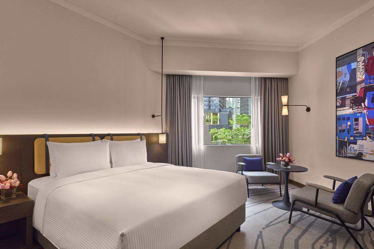 Habitación de hotel con una cama grande, una silla, una mesita y una ventana con vistas a edificios y vegetación.