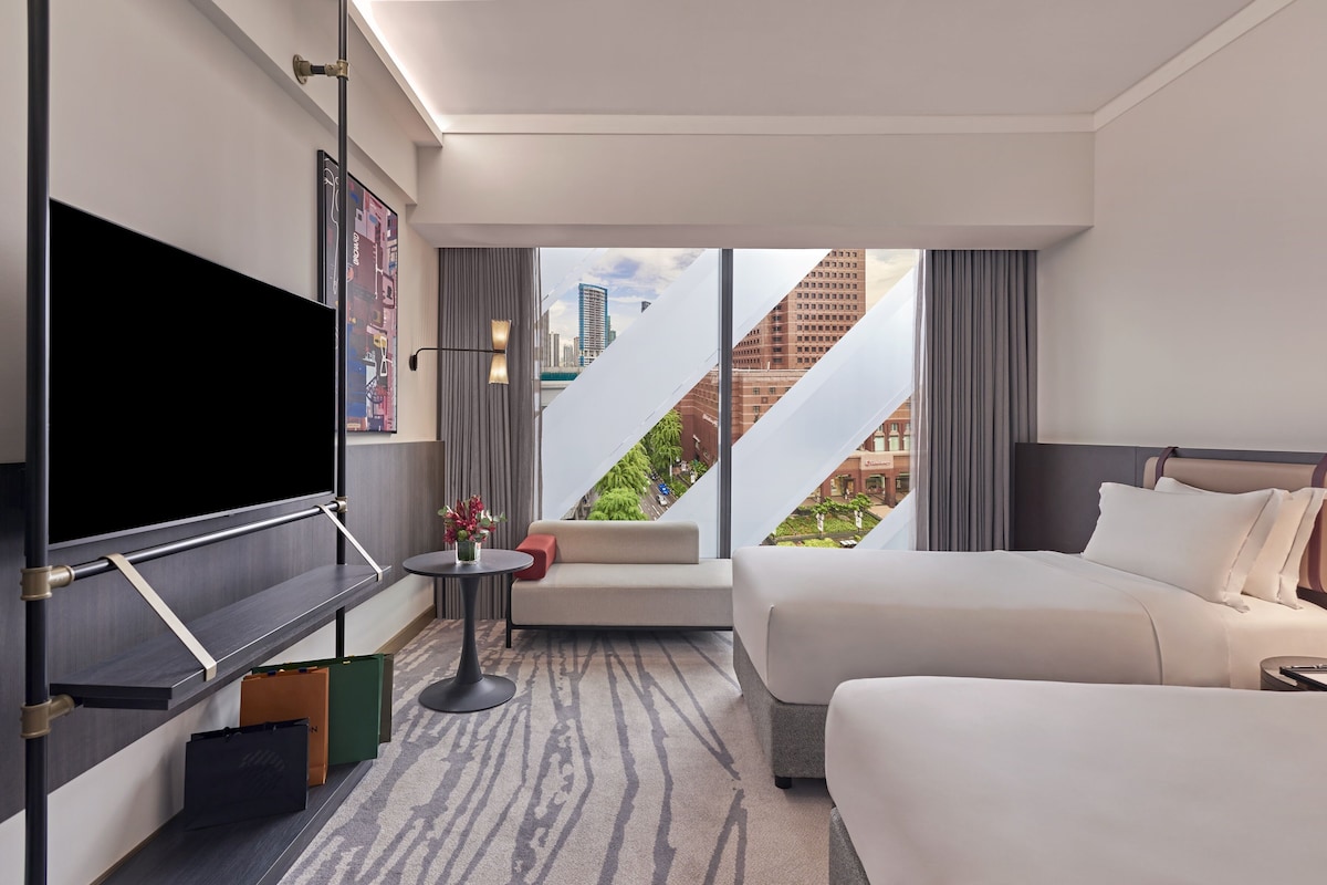 Una habitación de hotel moderna con una cama grande, un escritorio, un televisor de pantalla plana, una mesita con un jarrón y vistas a la ciudad a través de amplios ventanales.