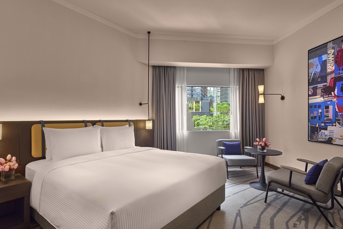 Habitación de hotel con una cama grande, una silla, una mesita y una ventana con vistas a edificios y vegetación.