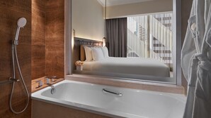 A modern bathroom with a bathtub, shower, and a large mirror reflecting a bedroom with a bed and curtains.