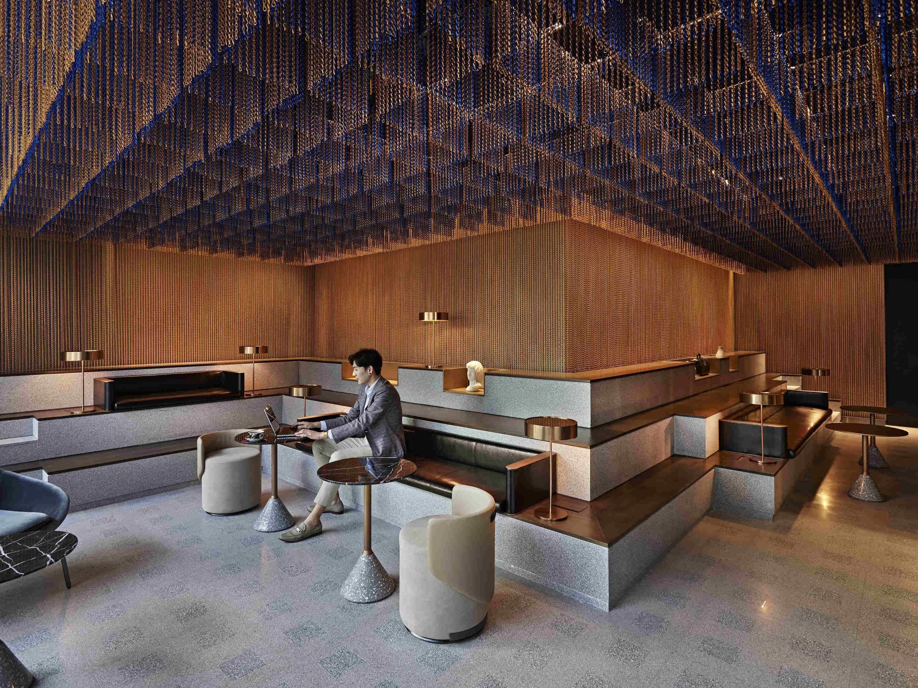 A modern lobby with a man working on a laptop, featuring a unique ceiling design, seating areas, and a reception desk.