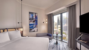 A hotel room with a large bed, a sofa, a small table, a TV, and a city view through the window.