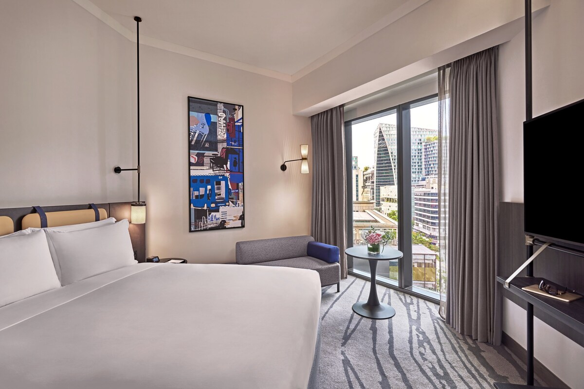 Habitación de hotel con una cama grande, un sofá, una mesita, un televisor y vistas a la ciudad por la ventana.