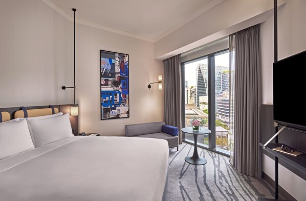 Habitación de hotel con una cama grande, un sofá, una mesita, un televisor y vistas a la ciudad por la ventana.