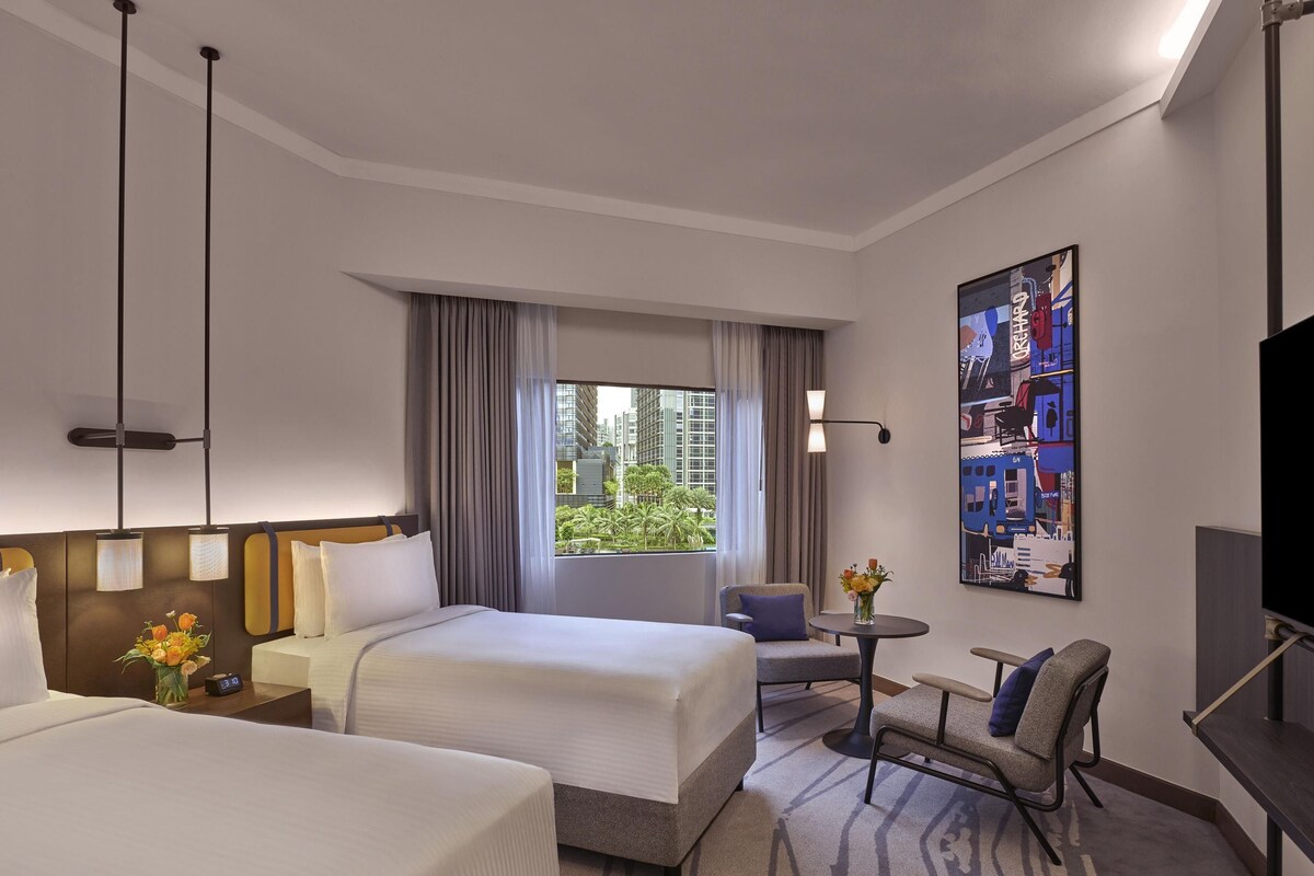 Habitación de hotel con dos camas, un escritorio, una silla, un televisor y un amplio ventanal con cortinas.