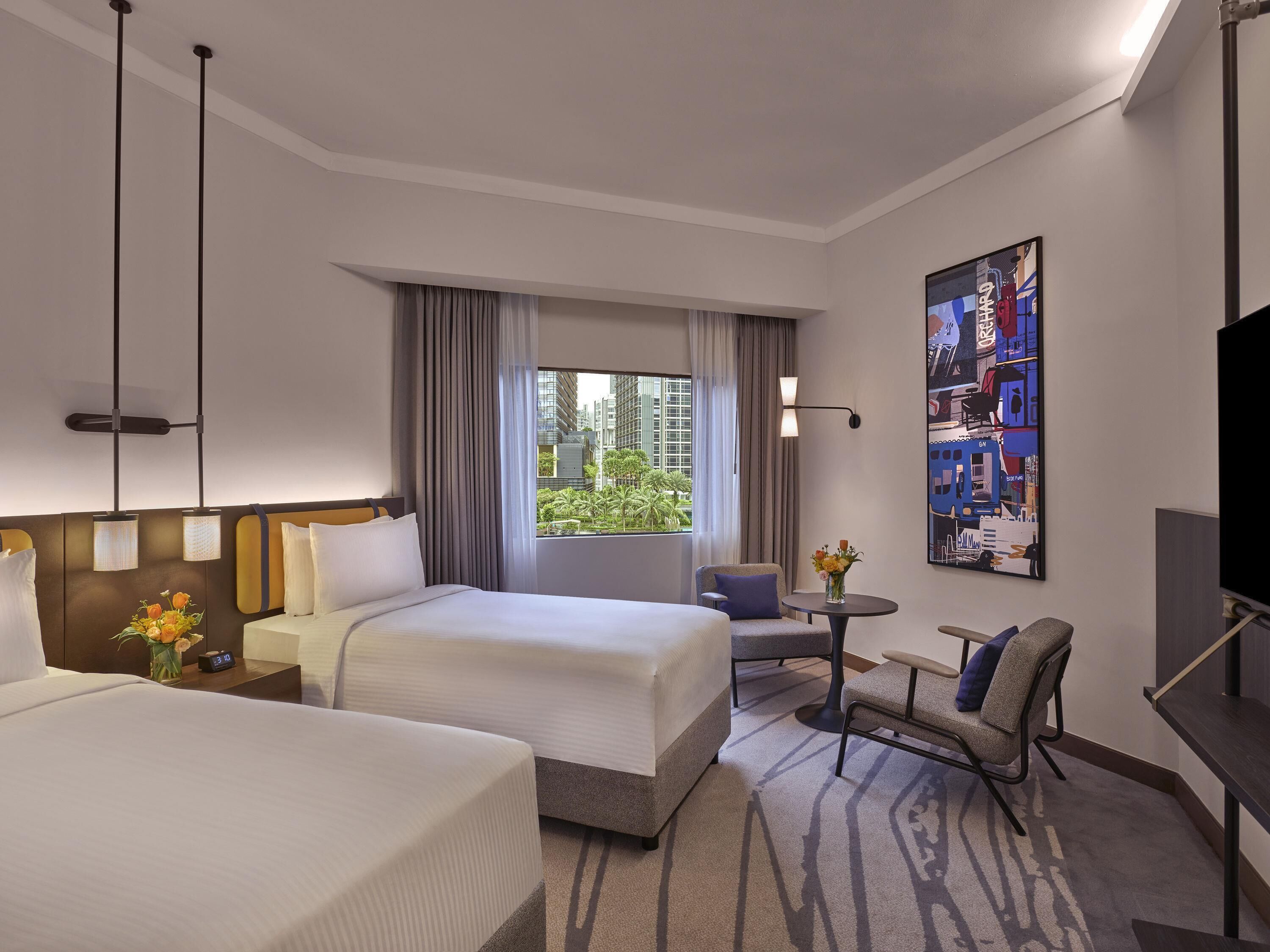 Ein Hotelzimmer mit zwei Betten, einem Schreibtisch, einem Sessel, einem Fernseher und einem großen Fenster mit Vorhängen.