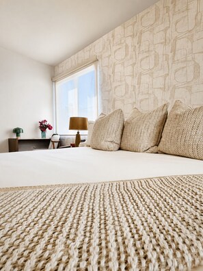 Un dormitorio con una cama grande, una ventana, una mesita de noche, una lámpara y un jarrón con flores.
