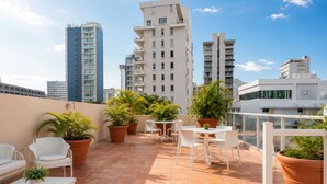 A rooftop terrace with potted plants, white outdoor furniture, and a cityscape view.