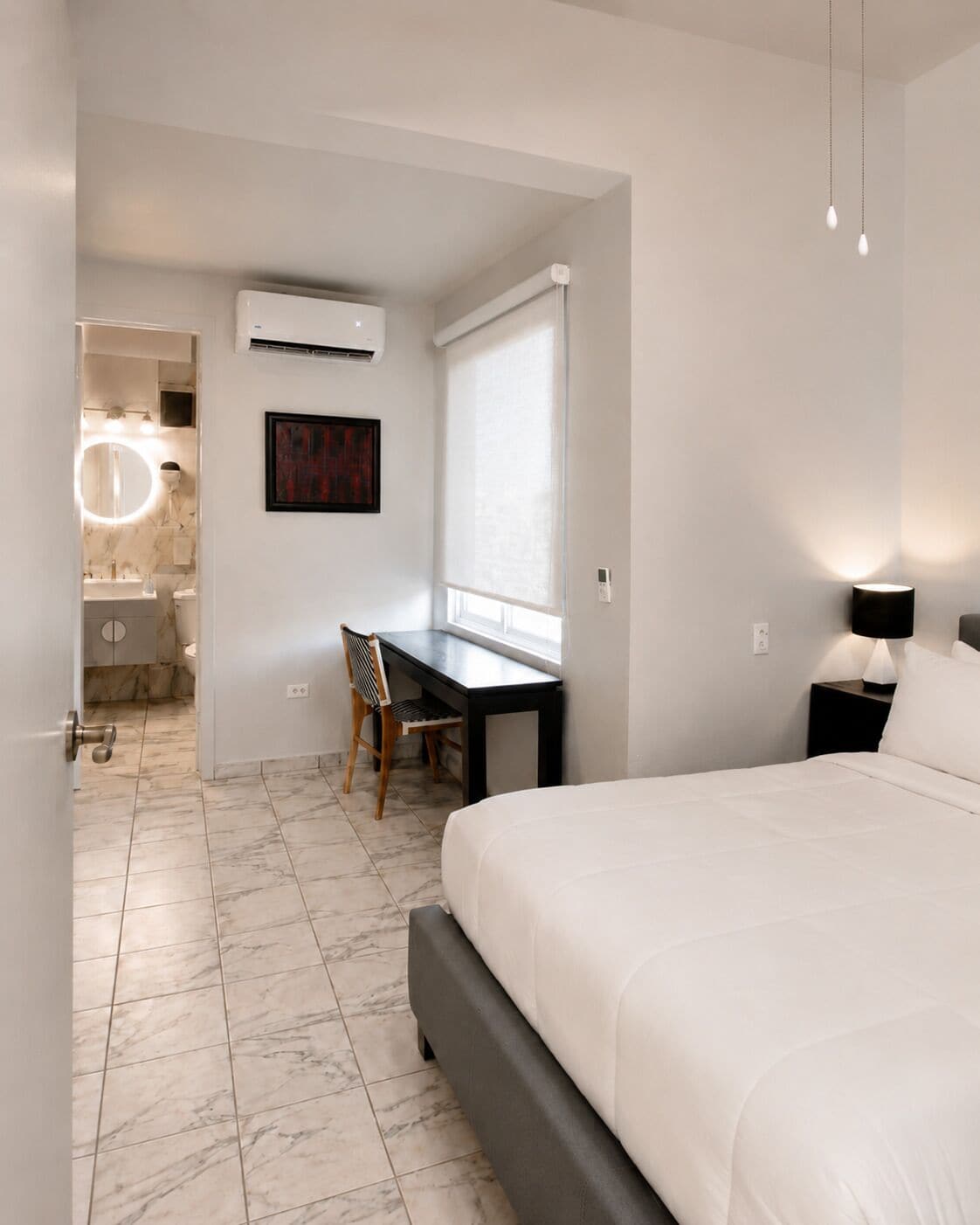 Un dormitorio con una cama, un escritorio y una silla. Se ve un baño por una puerta abierta.