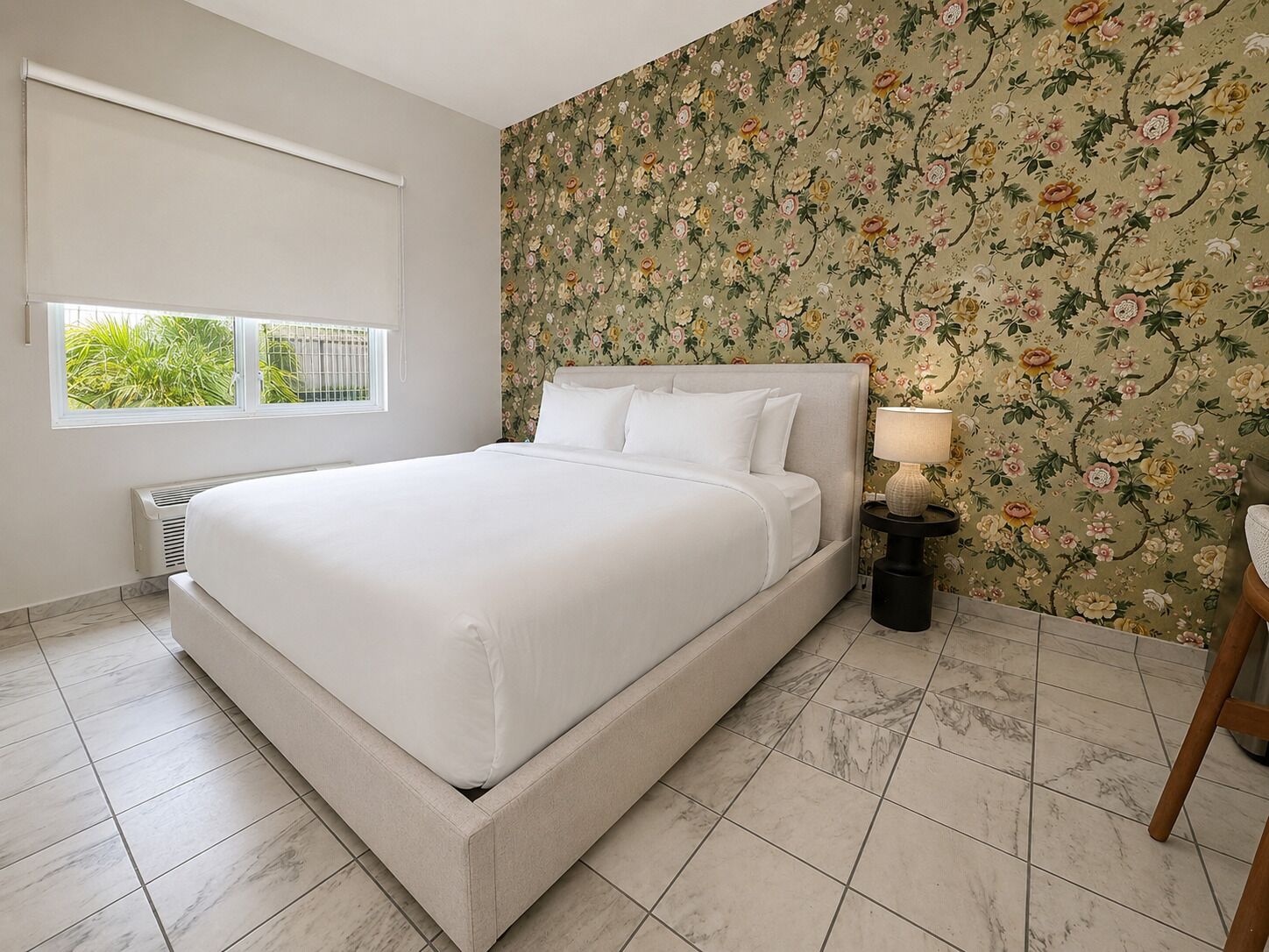 A bedroom with a bed, a window, a radiator, a nightstand with a lamp, and a floral wallpaper.