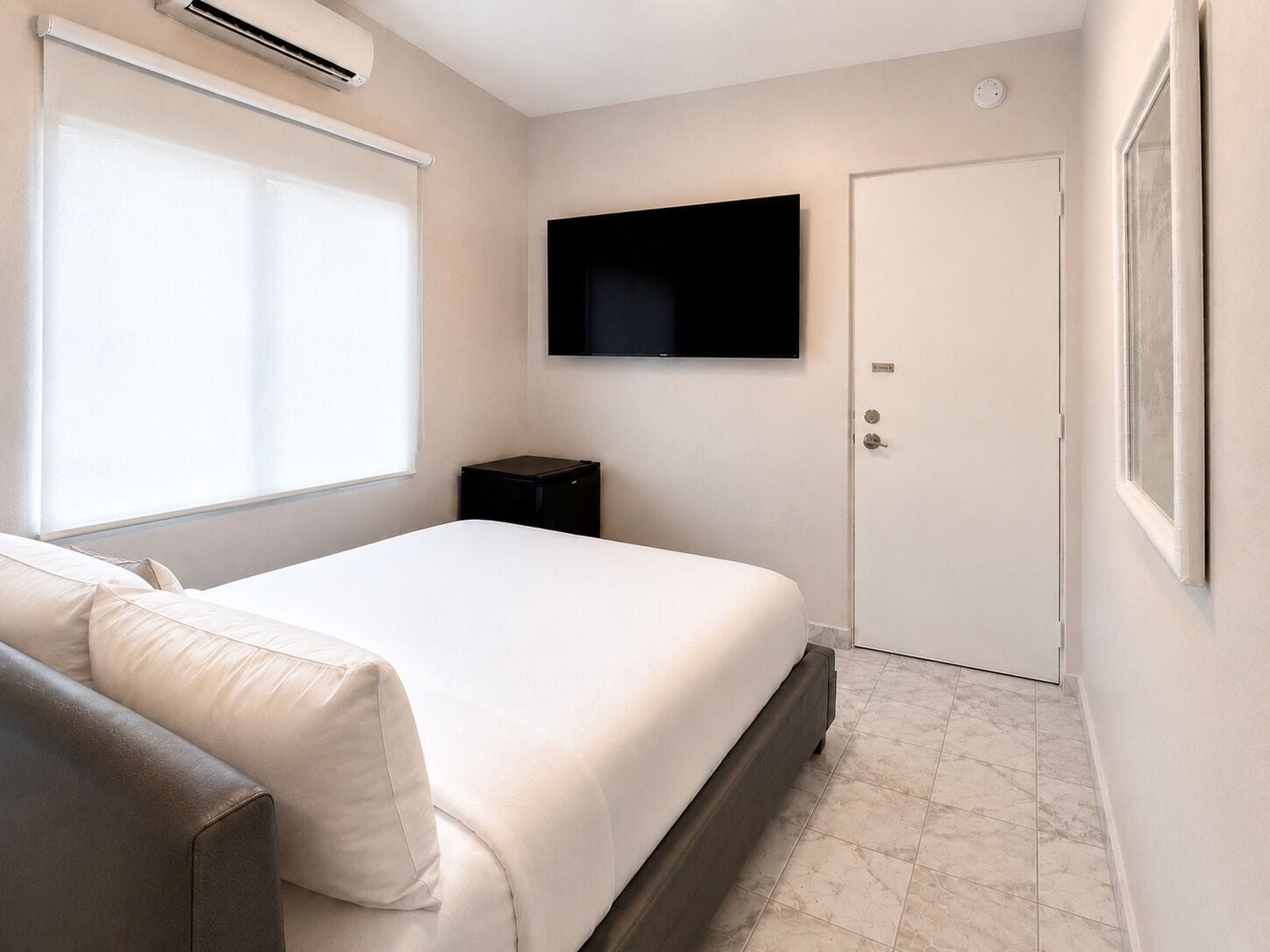 Ein Schlafzimmer mit einem Bett, einem an der Wand montierten Fernseher, einer Tür und einem Fenster mit Jalousien.