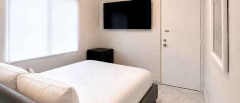 Ein Schlafzimmer mit einem Bett, einem an der Wand montierten Fernseher, einer Tür und einem Fenster mit Jalousien.