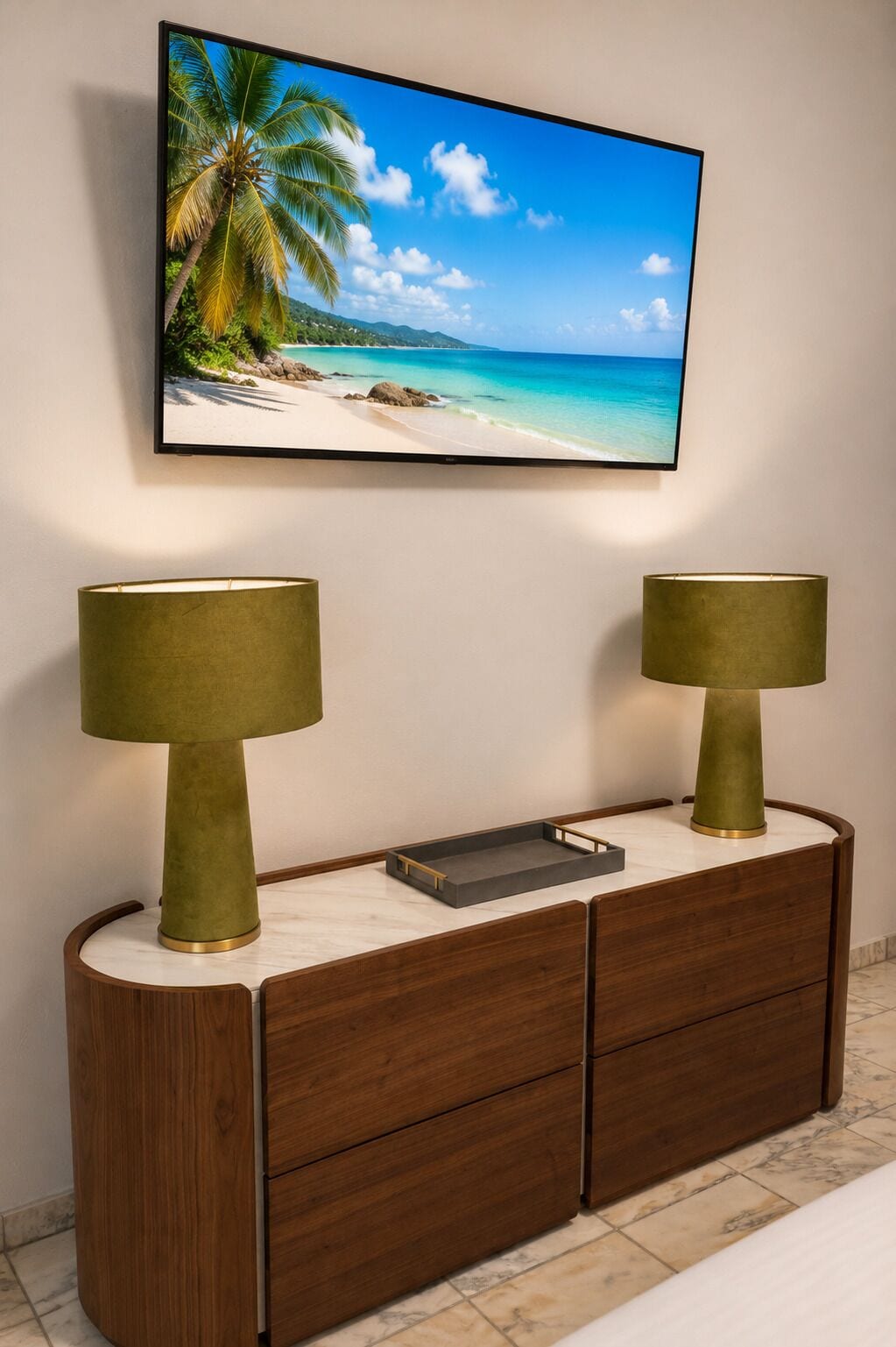 Un téléviseur fixé au mur, affichant une scène de plage tropicale, une console en bois avec des tiroirs et deux lampes dotées de pieds dorés.