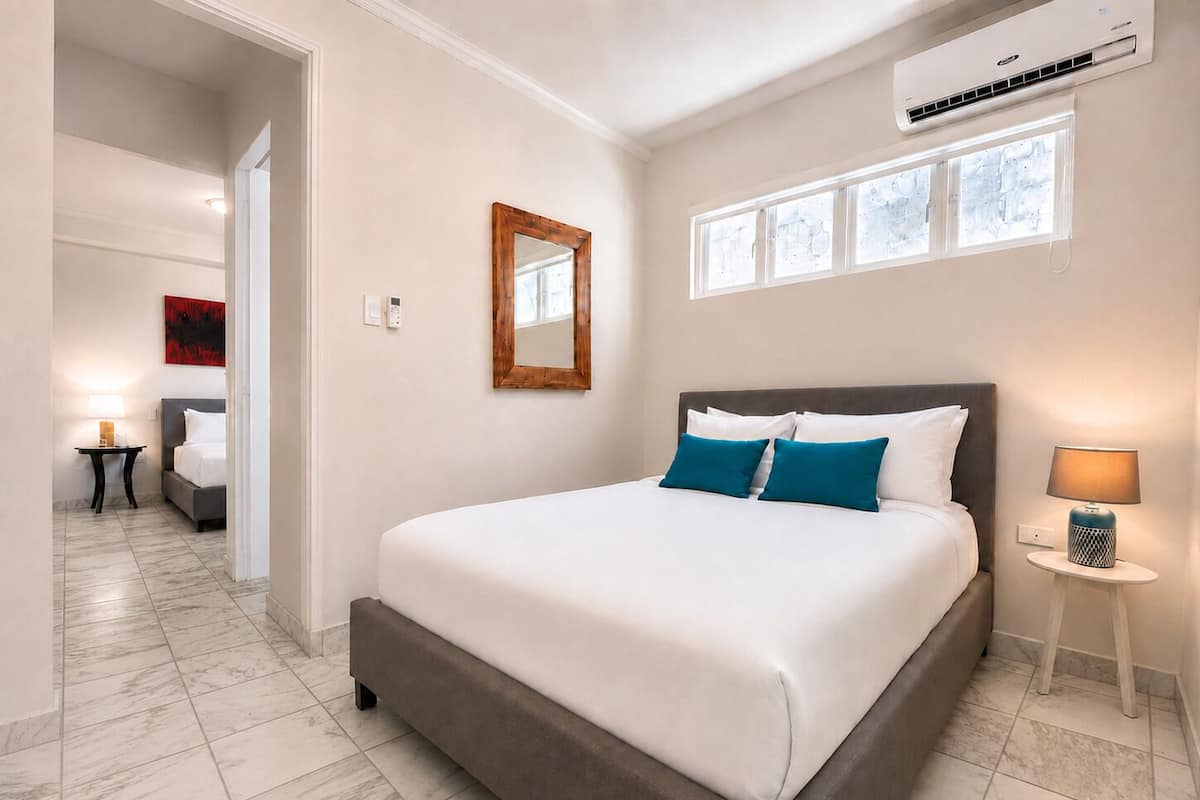 A hotel room with a large bed, a bedside table with a lamp, a window with blinds, and a wall-mounted air conditioning unit.