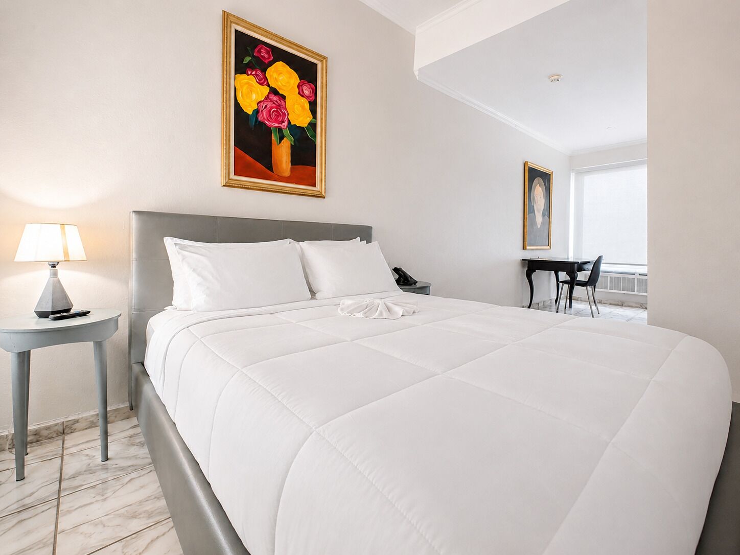 A hotel room with a bed, a bedside table with a lamp, a painting on the wall, and a desk with a chair.