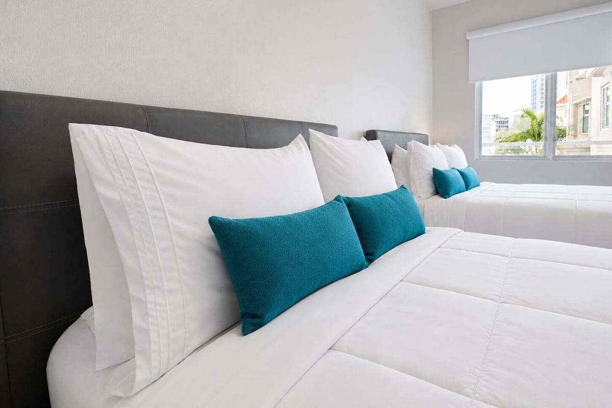 A hotel room with a large bed, two pillows, a window with a view of buildings, and a plain wall.