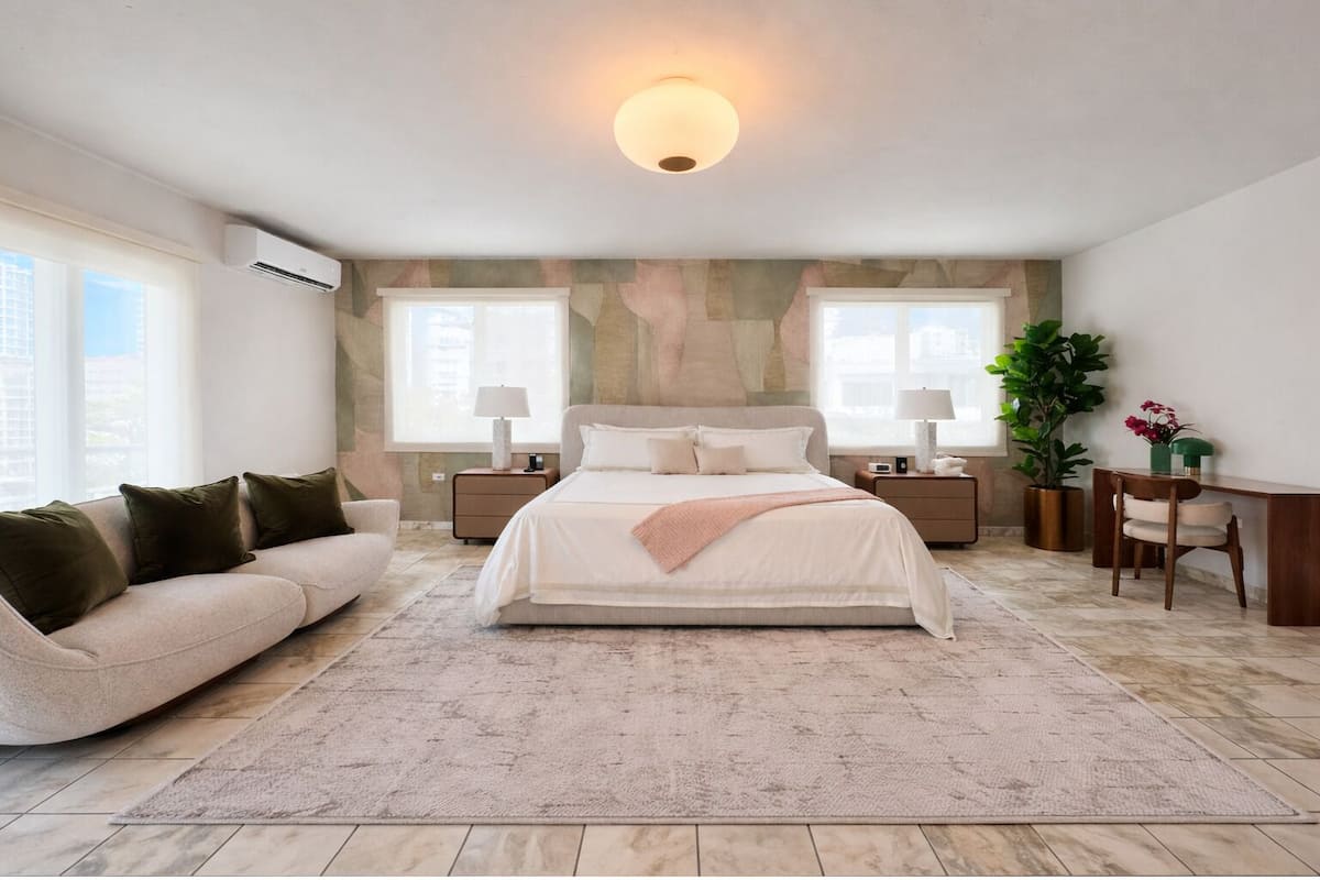 A bedroom with a bed, a sofa, a desk, a chair, a lamp, a potted plant, and a wall-mounted air conditioner.