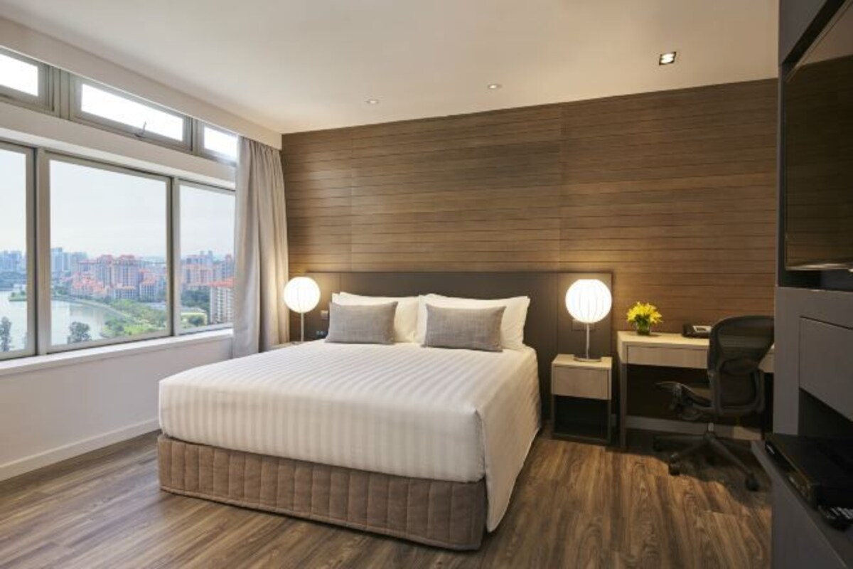 Ein modernes Hotelzimmer mit einem großen Bett, einem Schreibtisch und einem Sessel. Zwei Nachttischlampen und ein Blick auf die Stadt durch große Fenster.