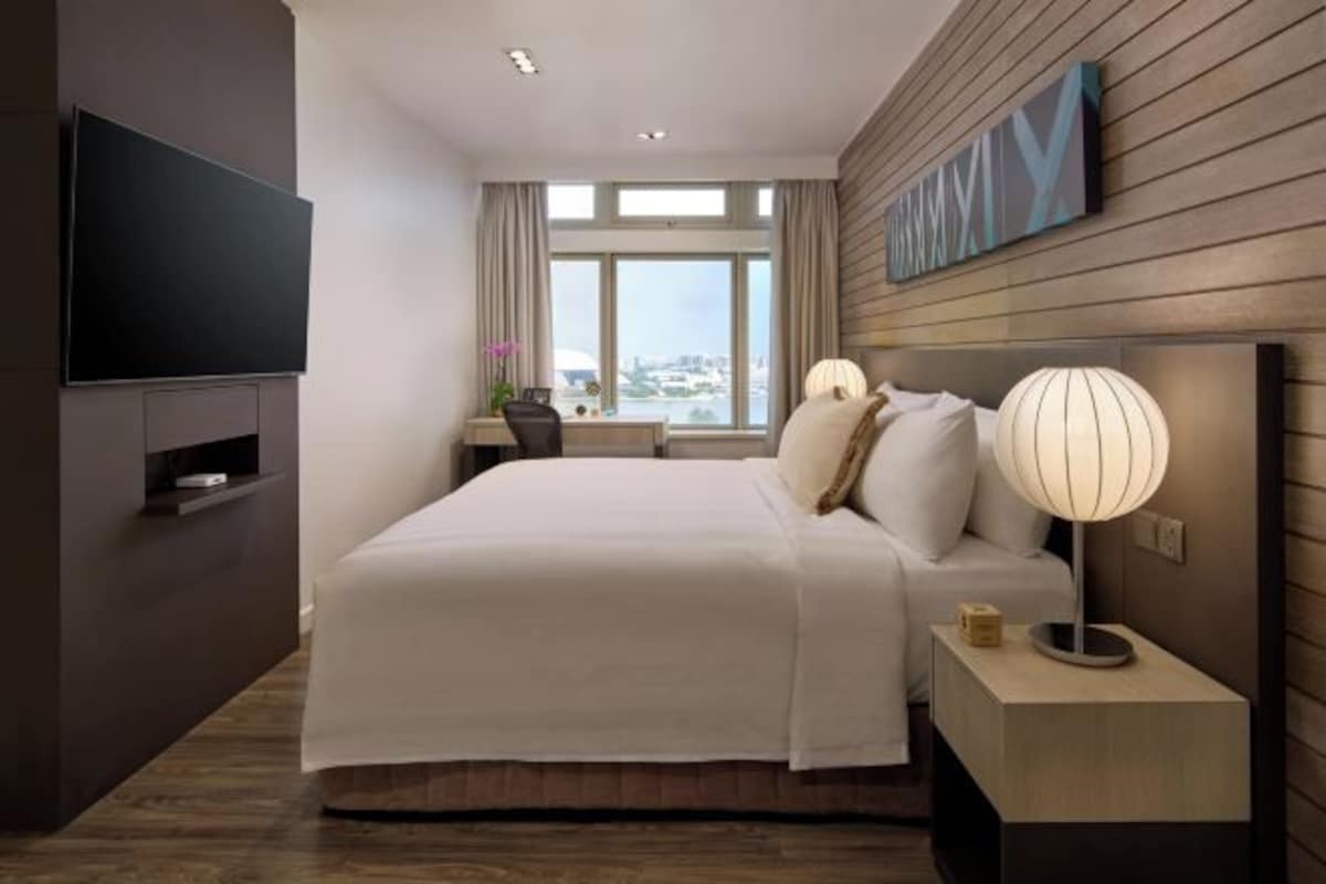 Ein modernes Hotelzimmer mit einem großen Bett, einem Flachbildfernseher, einem Nachttisch mit Lampe und einem Fenster mit Blick auf die Stadt.