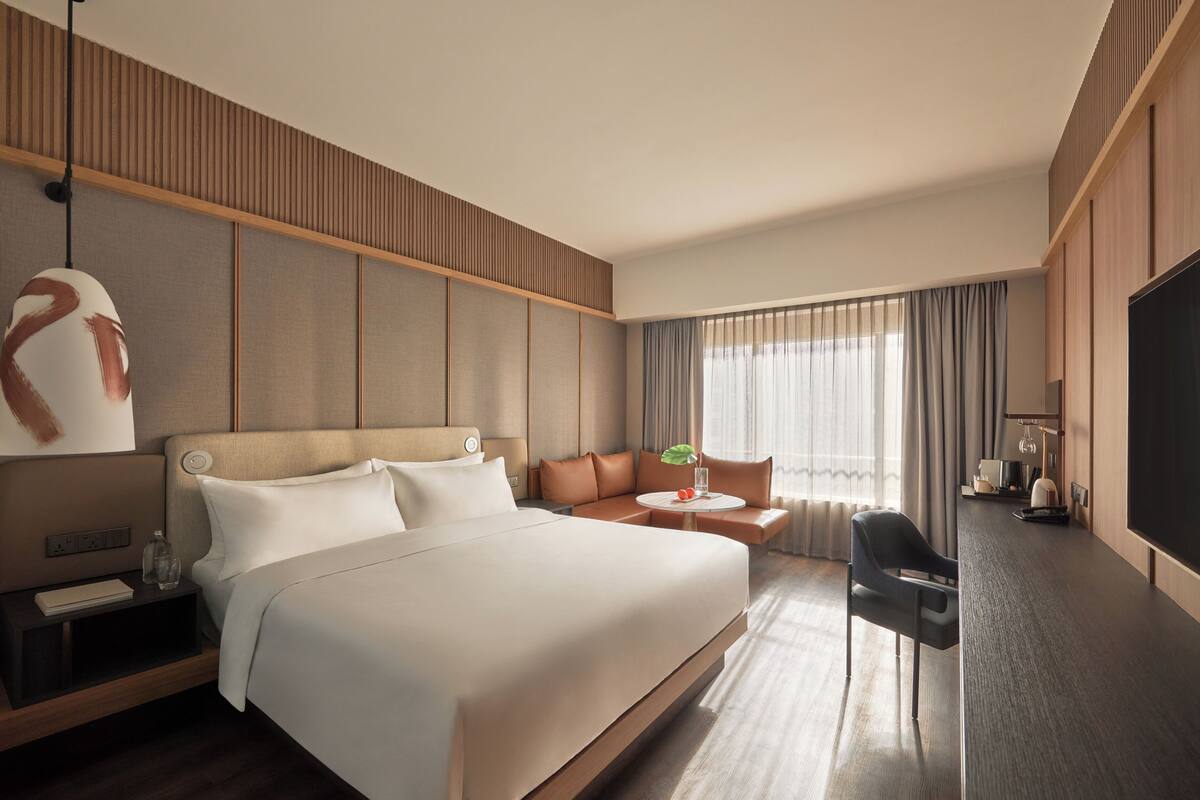 A modern hotel room with a large bed, a seating area with a sofa and armchairs, a flat-screen TV, and a large window with curtains.