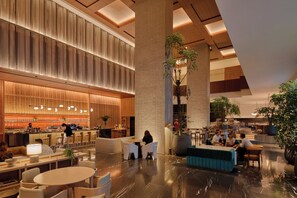 A modern hotel lobby with a bar area, seating arrangements, and large potted plants.