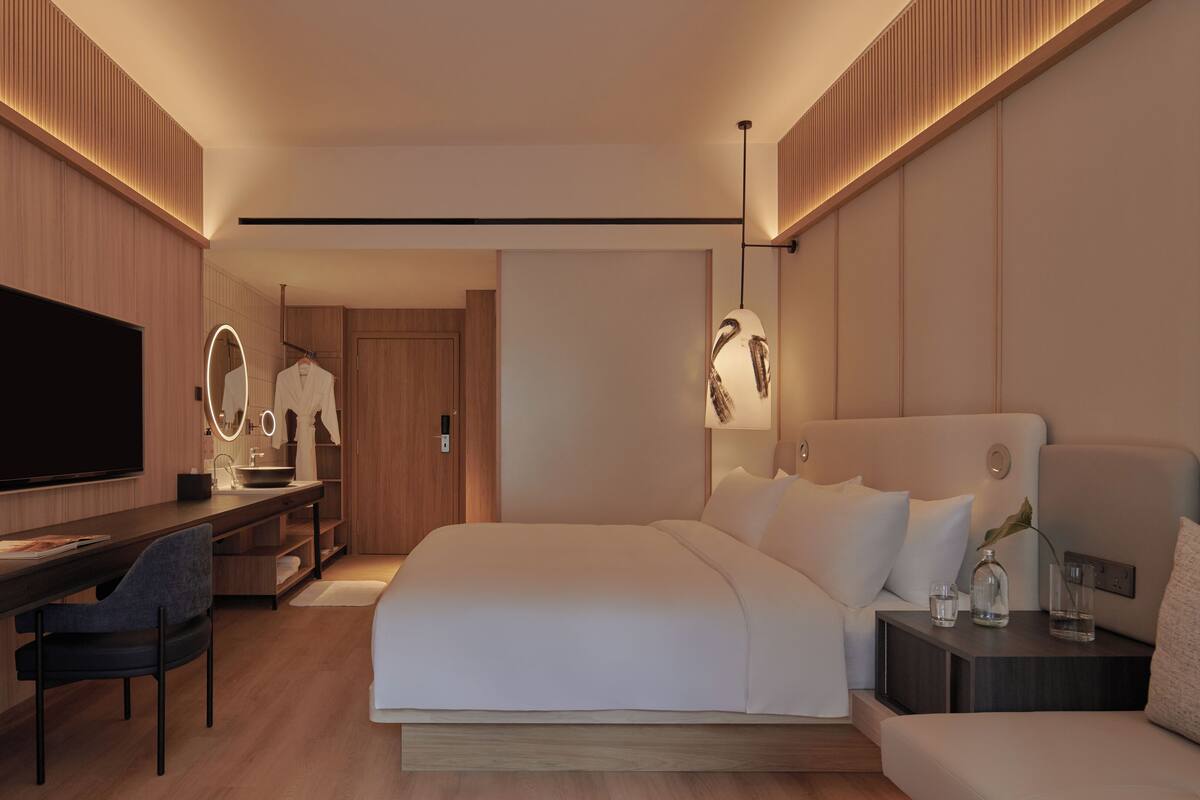 A modern hotel room with a large bed, a desk with a flat-screen TV, a vanity with a mirror, and a bedside table with a lamp.