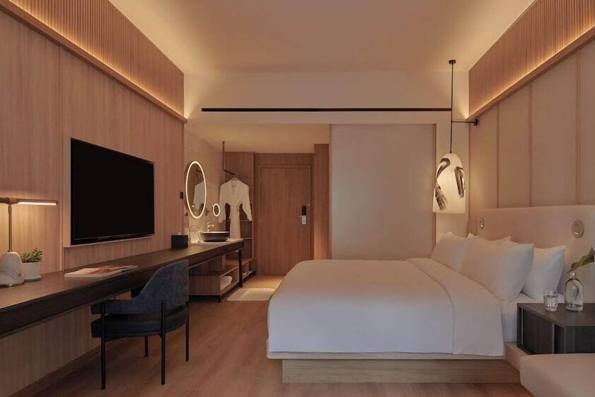 A modern hotel room with a large bed, a desk with a flat-screen TV, a vanity with a mirror, and a bedside table with a lamp.