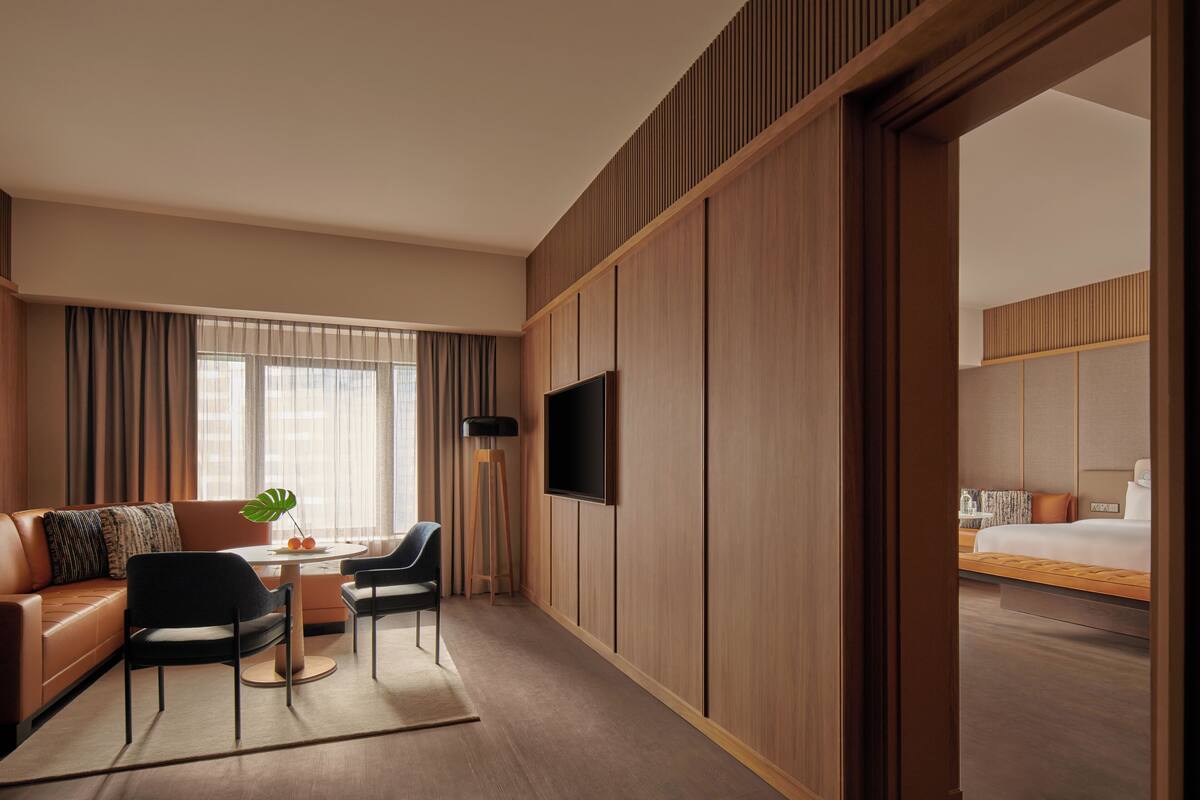A modern hotel room with a wooden panel wall, a flat-screen TV mounted on the wall, a sofa, a dining table with chairs, and a bed with a wooden headboard.