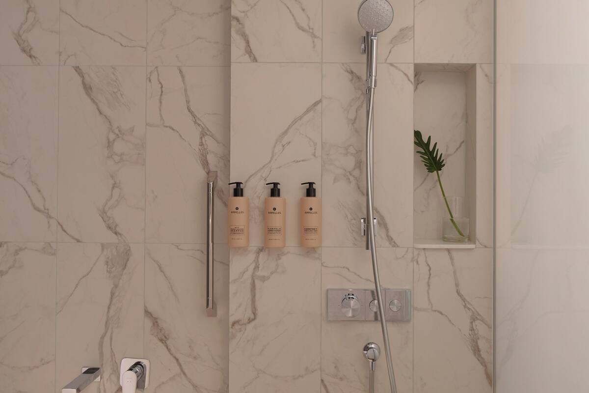 A modern bathroom with a marble wall, a freestanding bathtub, a handheld showerhead, and three toiletry bottles on a shelf.