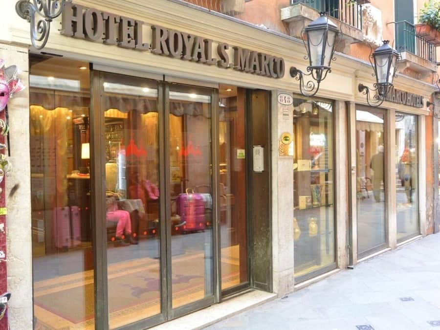 Exterior do HOTEL ROYAL S MARCO, com grandes portas de vidro, fachada clássica e uma placa com o nome do hotel.