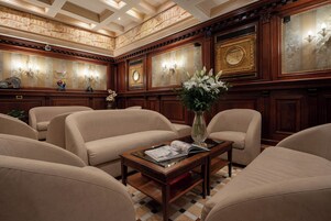 Quarto luxuoso com amplas sofás bege, mesa de centro de madeira com um vaso de flores e decoração requintada nas paredes, incluindo espelhos e obras de arte emolduradas.