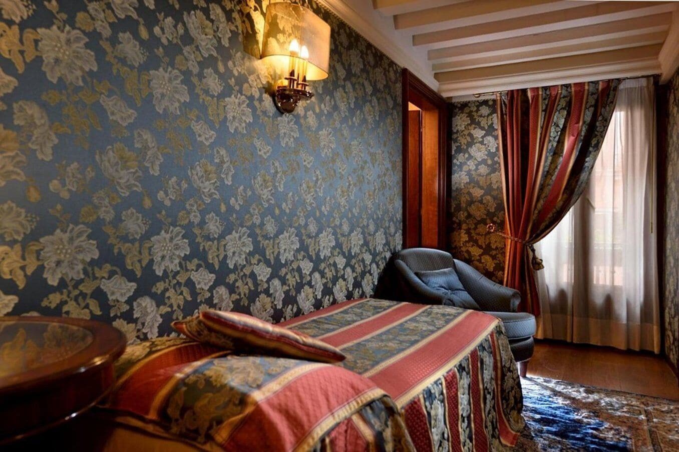Una habitación con papel tapiz estampado, una cama con manta roja y azul, una silla y una lámpara.