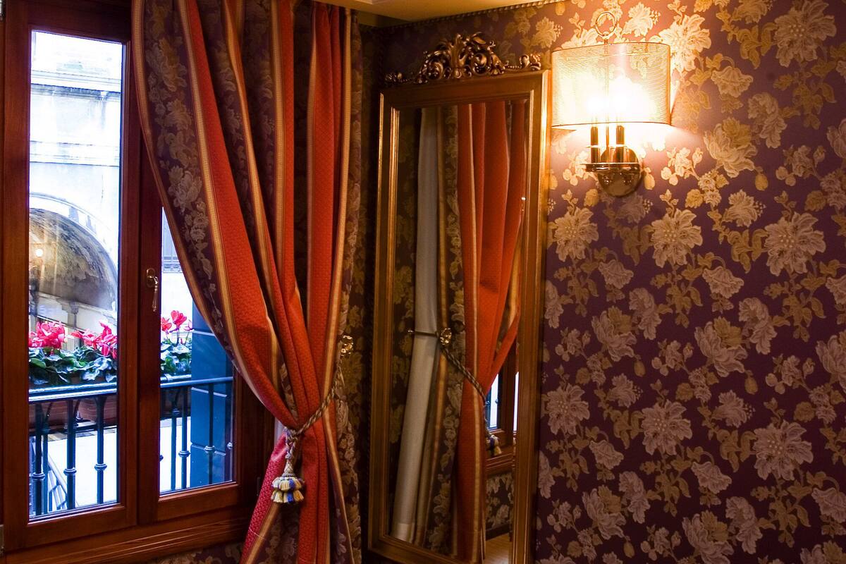 A room with a bed, curtains, a wall-mounted lamp, and a window with a view of flowers outside.