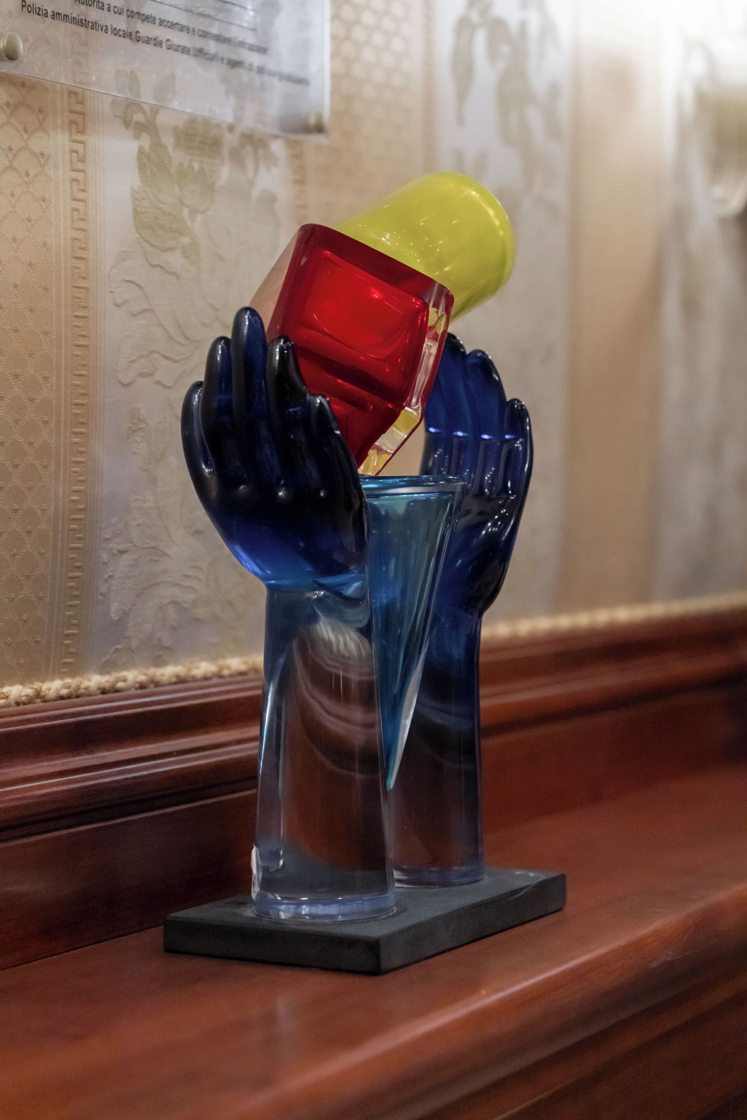 Una escultura de cristal decorativa que muestra manos sosteniendo una copa y un objeto rojo, sobre una superficie de madera.